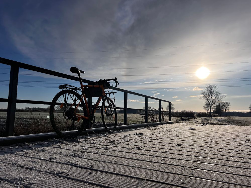 Fahrrad lehnt am Metallgeländer einer mit Frost überzogenen Brücke vor tief stehender Wintersonne