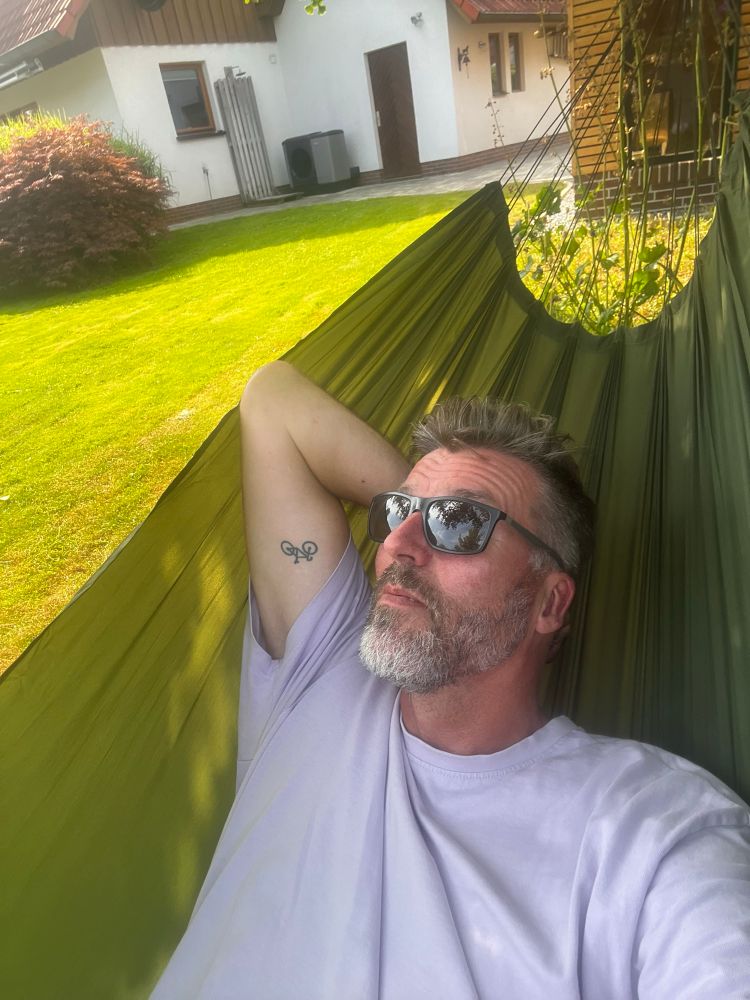 Mann in einer grünen Outdoor-Hängematte mit Sonnenbrille. Im Hintergrund ein weiß verputztes Haus