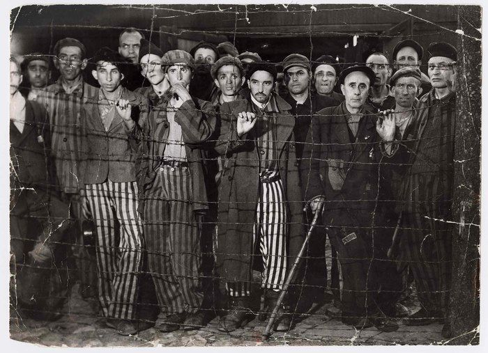 A group of prisoners, some in stripped uniforms, others in ragged clothing, look toward the photographer from behind a wire fence. Photograph by Margaret Bourke-White, Life Magazine, 1945.