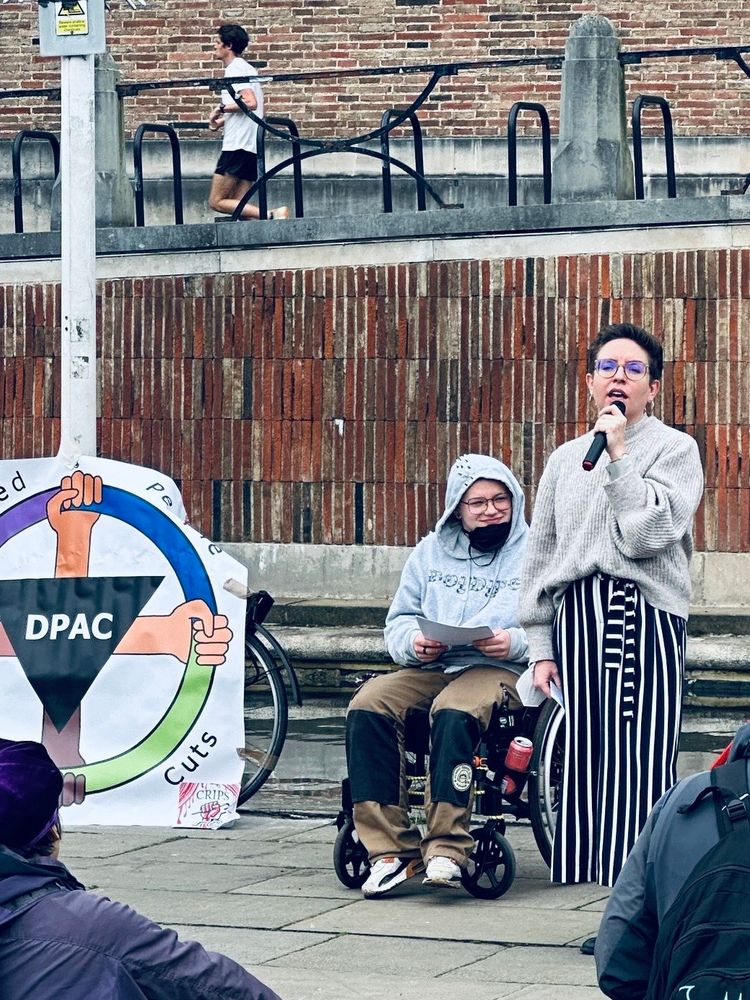 Green Party co-leader Carla Denyer speaking holding a mic, next to a protest organiser and Disabled People Against Cuts banner 