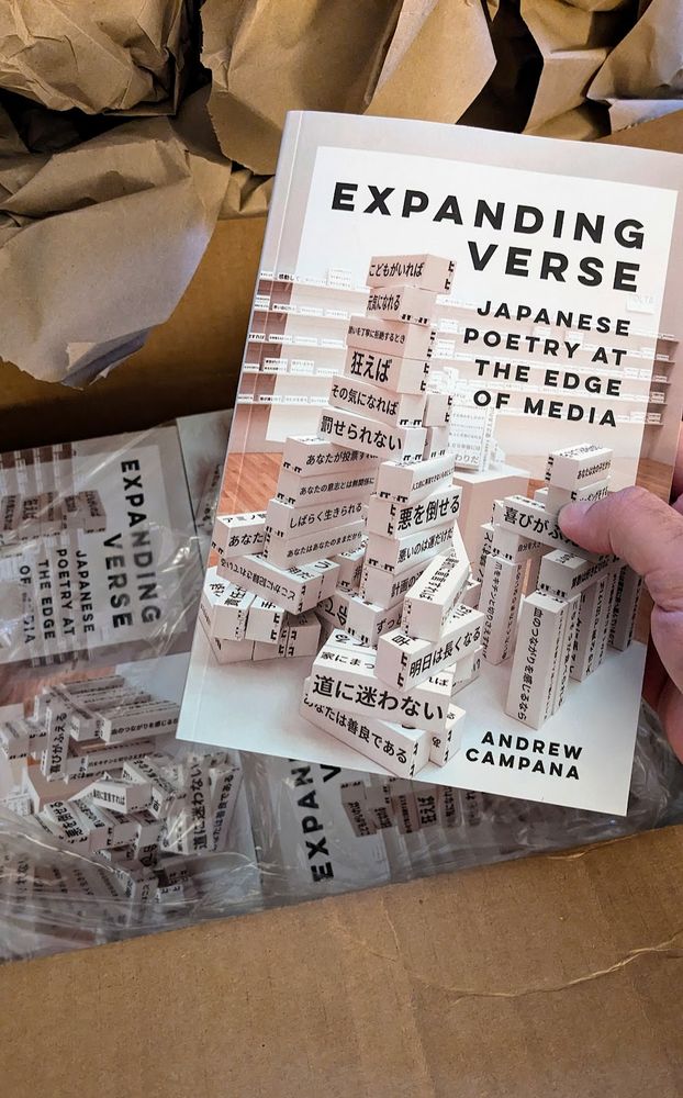 The author's hand picks up a copy of the book Expanding Verse from a just-opened cardboard box filled with other copies of the same book. 