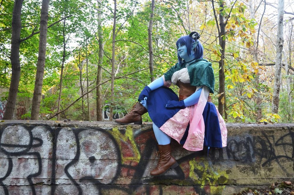 Embyr dressed as Jester Lavorre from Critical role in a blue and pink skirt and green cape-let sitting on a low stone wall in the middle of the woods  