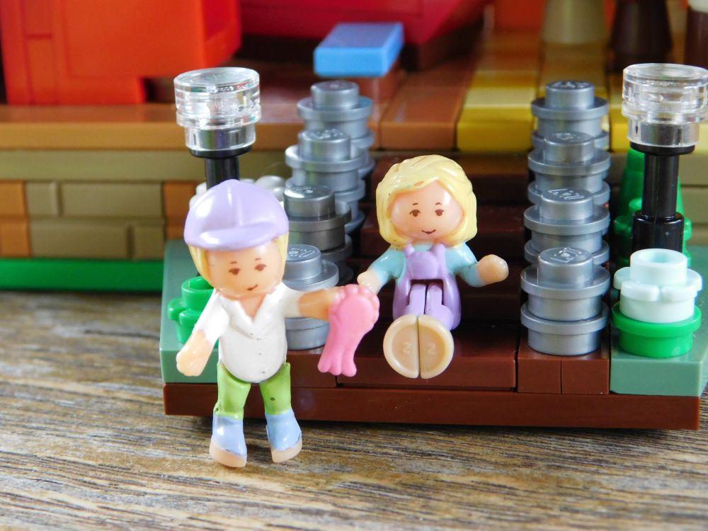 the two Polly Pockets outside their Lego dollhouse, arms raised as if waving farewell to the viewer. the front of the dollhouse has brown stairs flanked by flowers and two tall black outdoor lamps