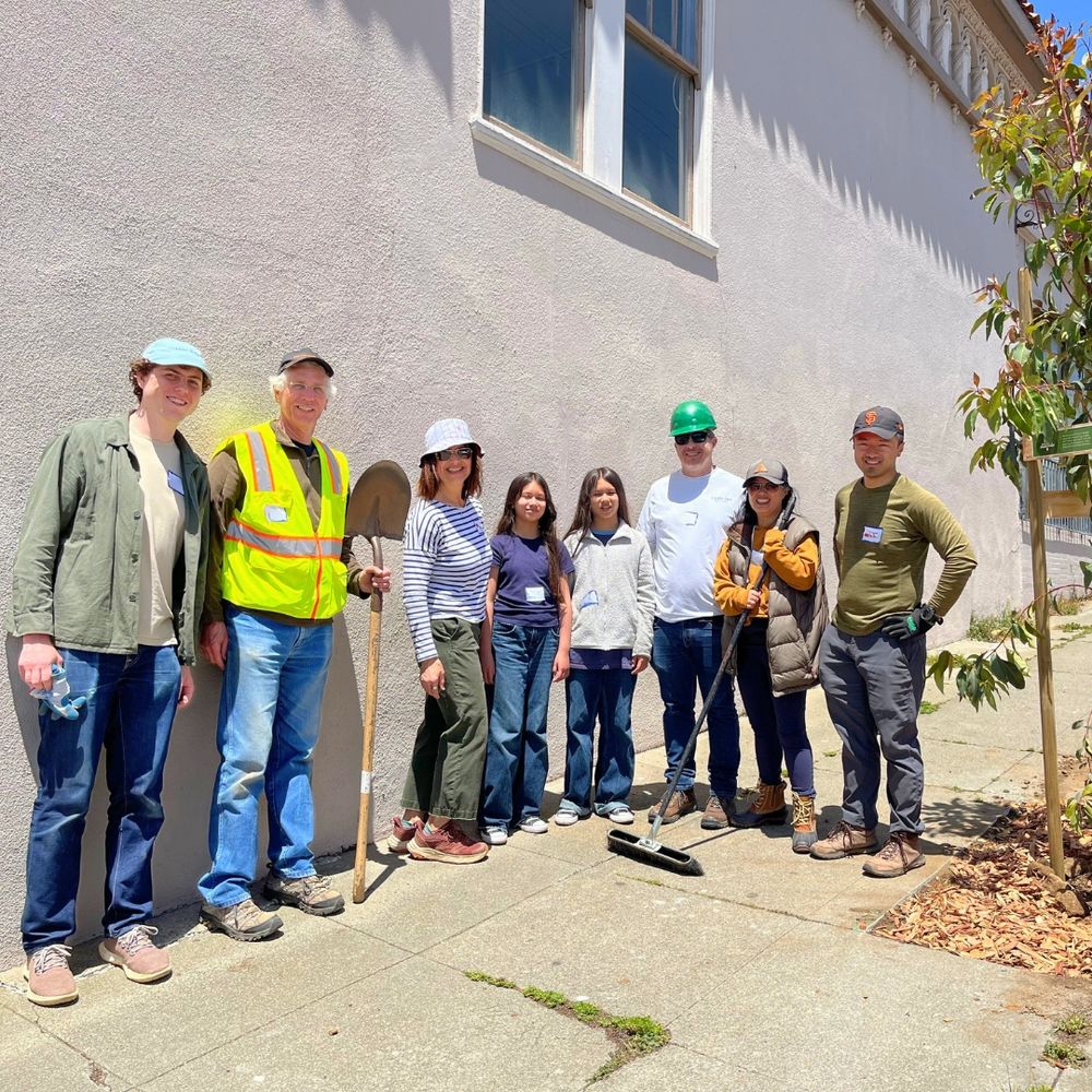 Eight smiling people stand near a newly planted tree.
