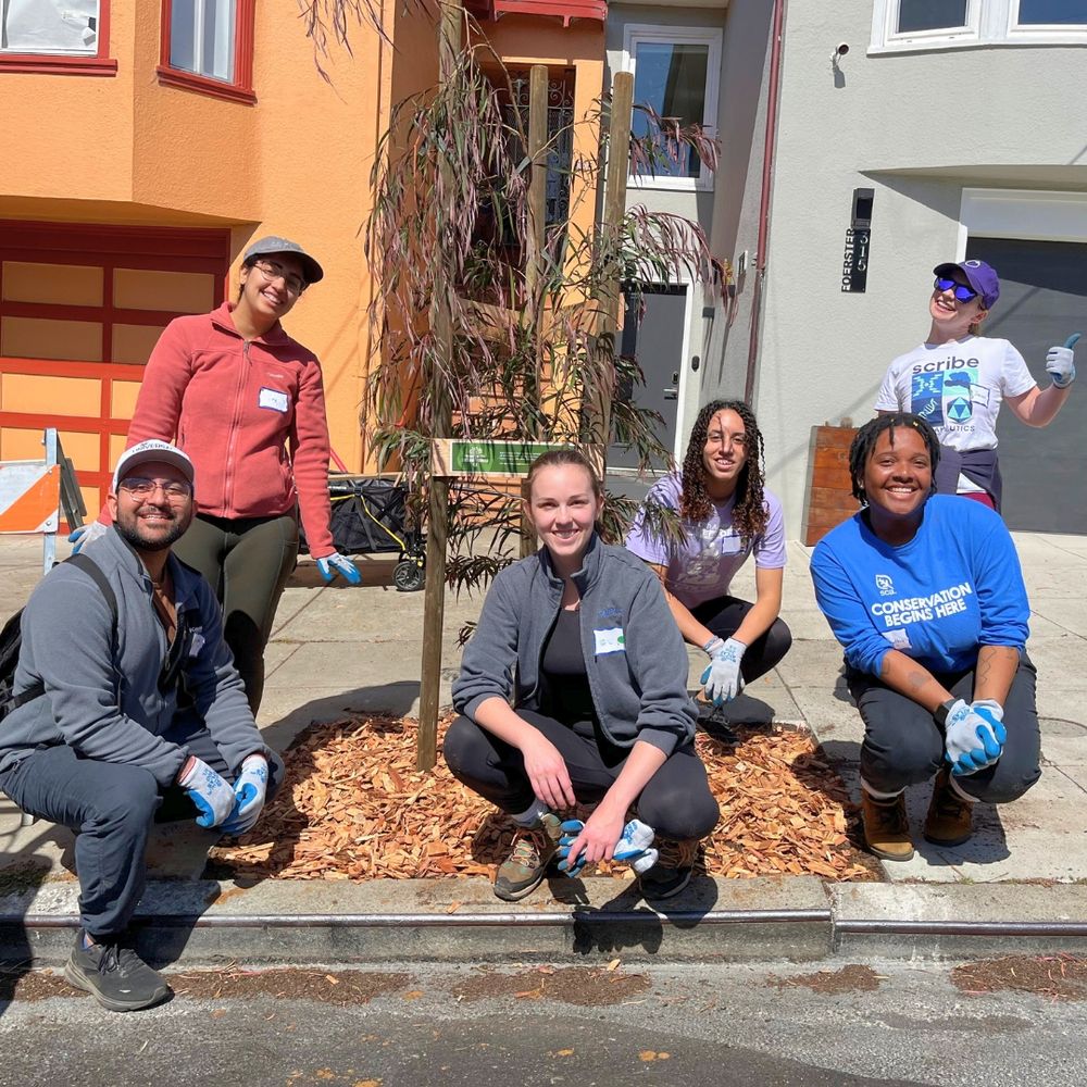 6 smiling volunteers and staff stand by a newly planted tree.