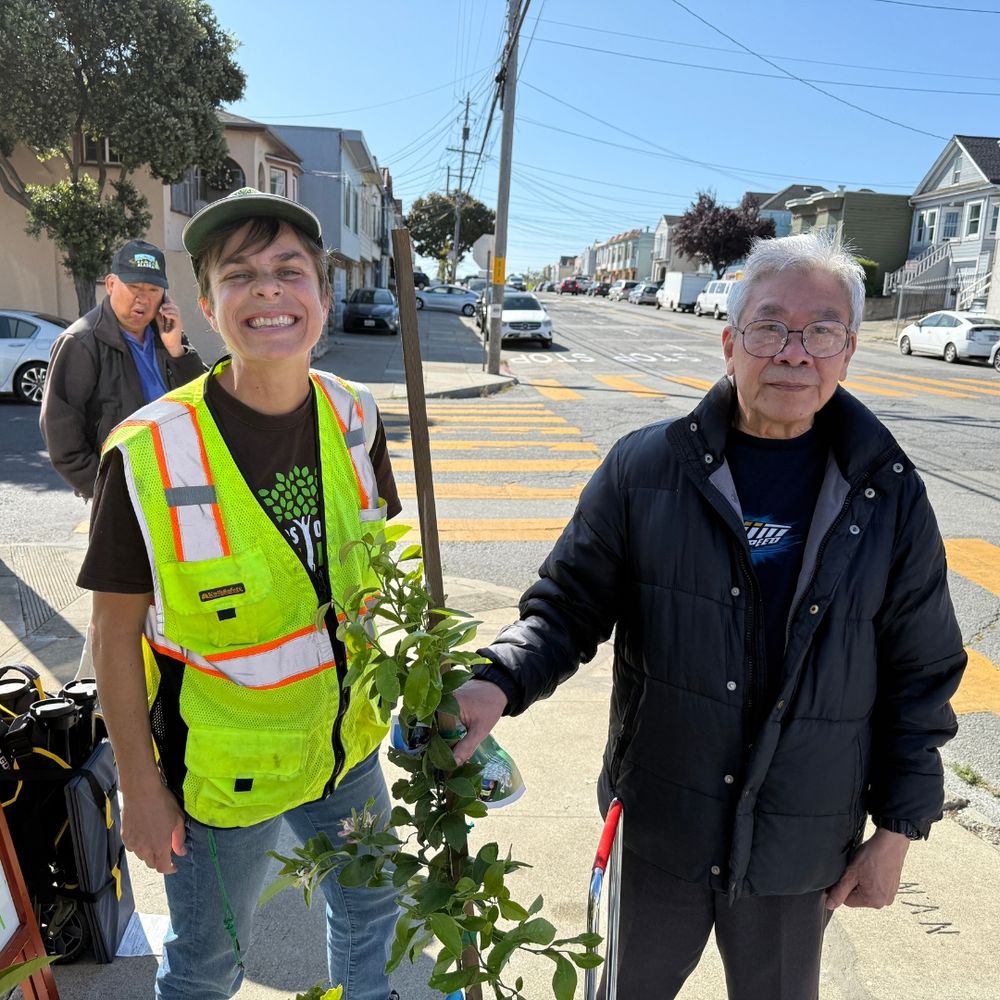 A Friends of the Urban Forest staff person and a smiling recipient of a lemon tree stand on either side of a lemon tree.
