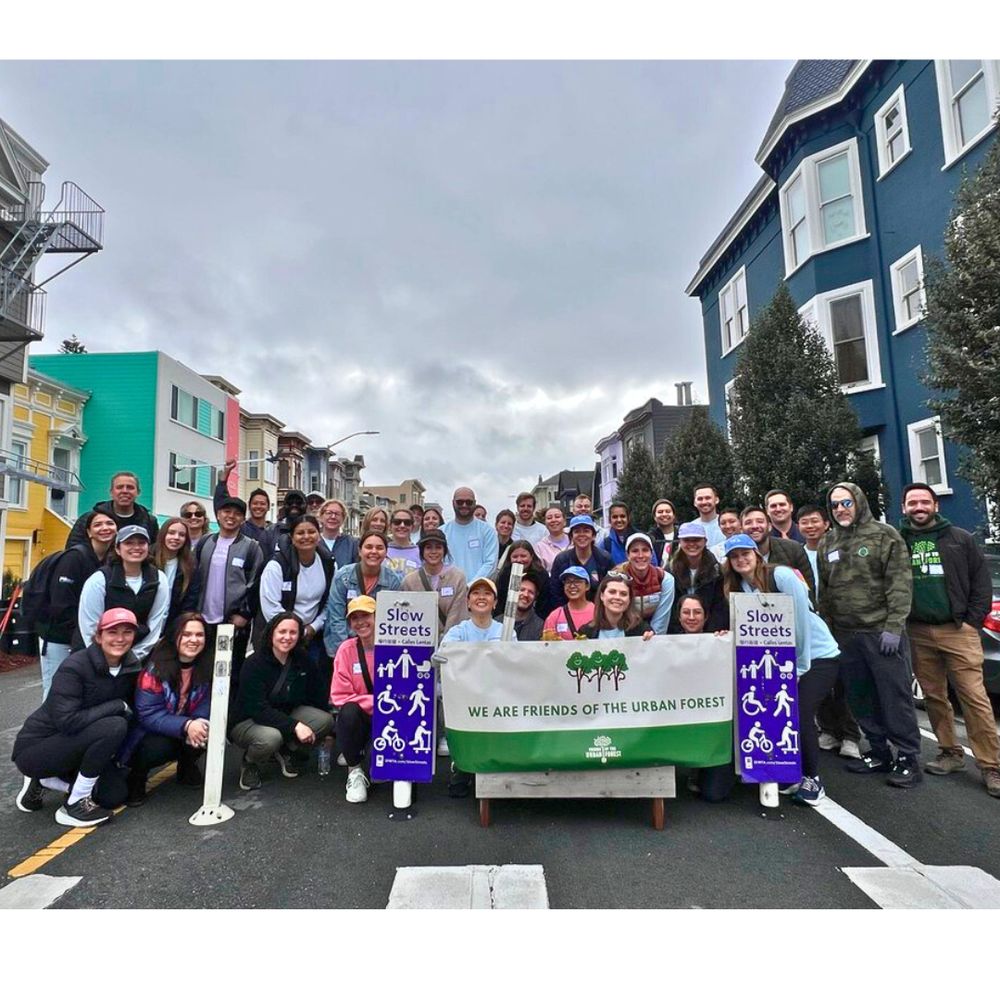 Group photo of 40+ volunteers and staff standing in a “slow street” and holding up a “We Are Friends of the Urban Forest” sign.