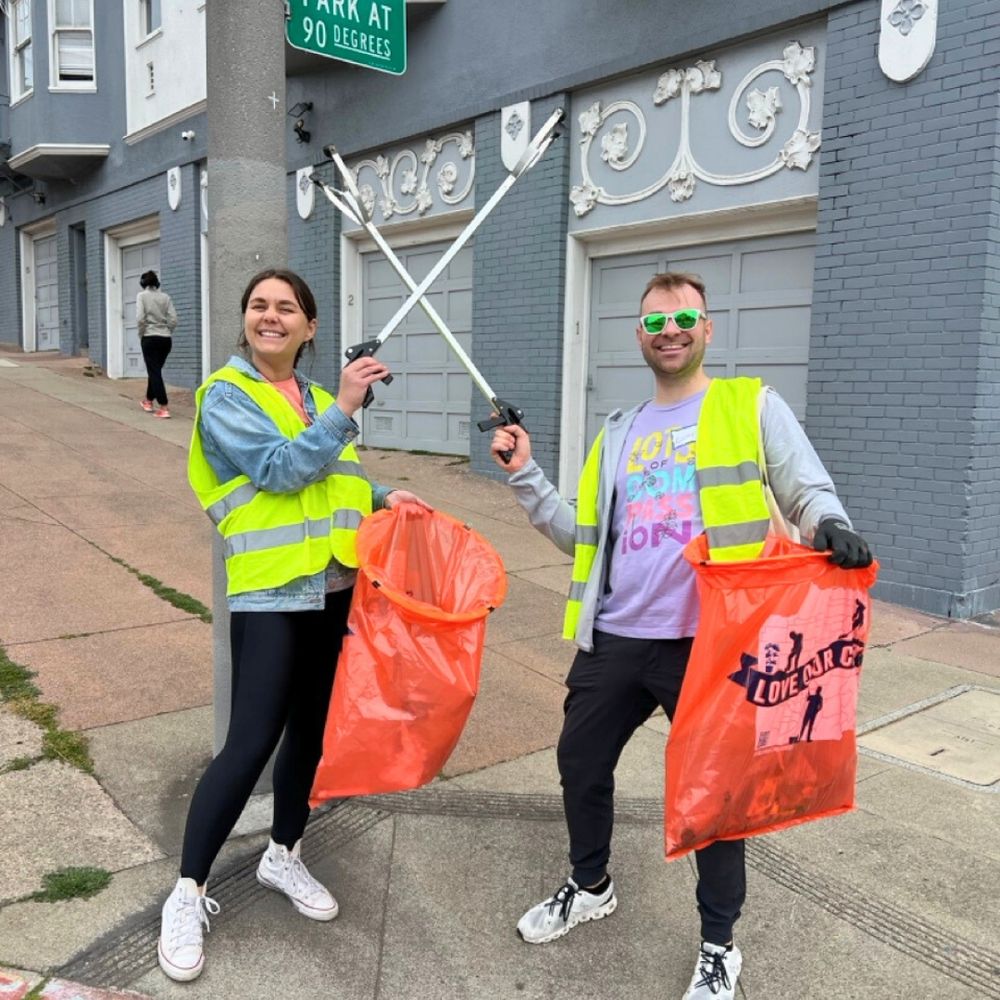 Two volunteers hold trash pickers in one hand and large orange trash bags in the other.