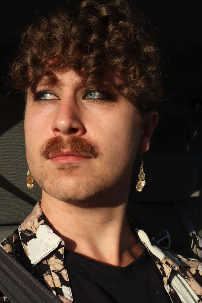A self portrait taken in the backseat of a car, with a very similar pose to the previous photo, but now my curls have puffed out a bit and cover my forehead more or less symmetrically, and there's fairly strong, warm lighting from the sun that has just begun to set. My head is turned just a litle more toward left-of-frame this time, and there's a strong shadow on the left side (right-of-frame) of my face and the background beyond it, with my left earring lit up in high contrast against it.