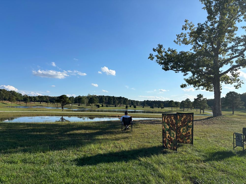 Another pond. Gorgeous blue sky with small puffy clouds on the horizon. A holding blind (where dog waits for tbh wide turn to run) is in the foreground. A handler is sitting in a chair waiting for his dog to come back from a long swim. 