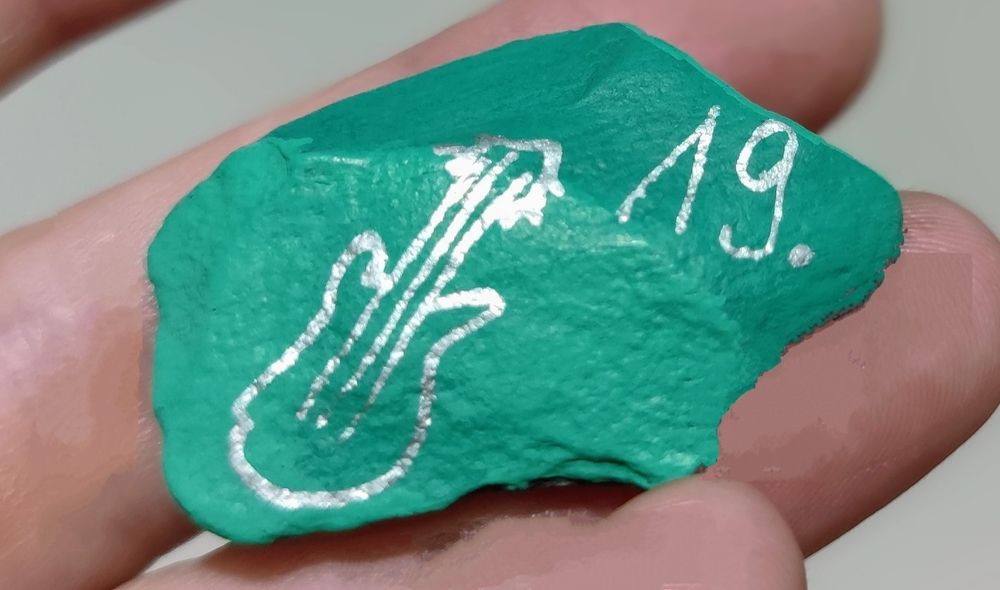 Bemalter grüner Stein, in Silber die Ziffer 19 und eine einfache Bassgitarre