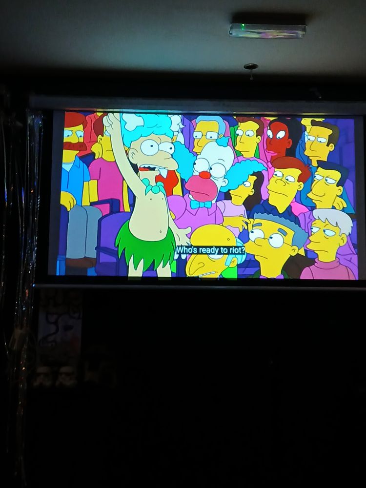Sideshow Mel stands up and yells in a vintage episode of "The Simpsons" (1989-Present). The episode is being shown on a television inside a restaurant that specialises with Chicken Wings.