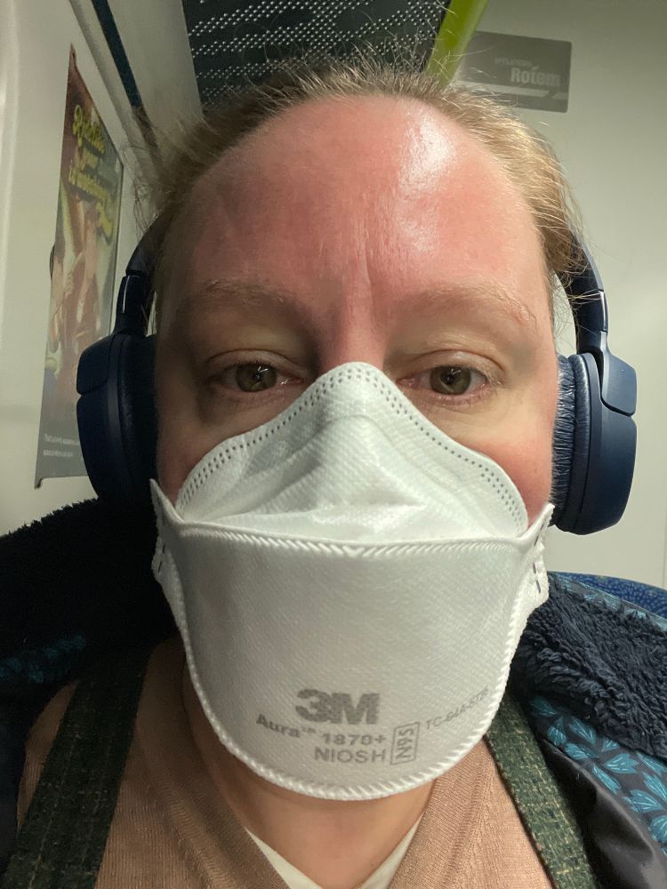 A white woman with green eyes and blue headphones wearing an Aura mask, sitting on a train seat. 