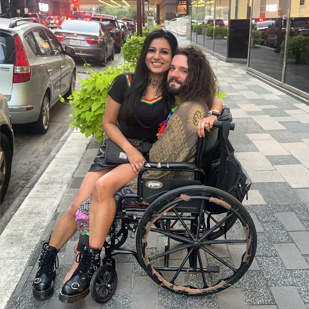 image shows a white man with long brown hair wearing a green pashmina in a wheelchair with a smiling brown woman sitting in his lap wearing a black top with a rainbow collar, a black leather skirt, and black boots in downtown houston 