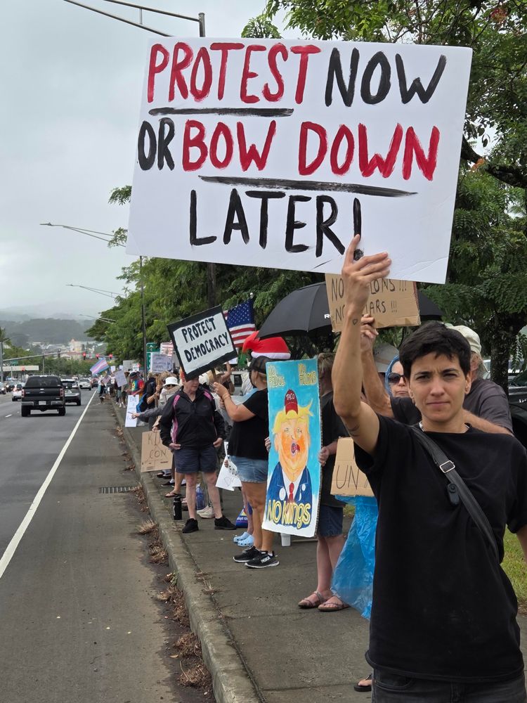 A line of protesters stretching down the sidewalk as far as you can see. A young woman in front olds a sign reading, "Protest Now or Bow Down Later!"