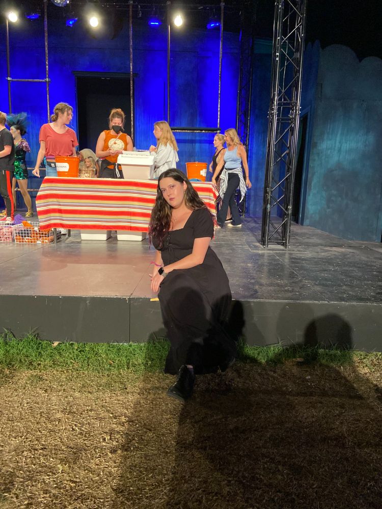 A person in a black dress with long brown hair sits on the lip of an outdoor stage. The wall behind them is deep blue and there is a picnic table loaded with merchandise on the stage.