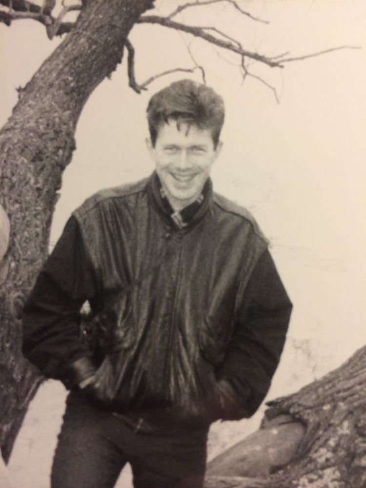 Monochrome photo of smiling Bill with 80s hair in a half-leather jacket. He’s by a lake side, facing the camera.