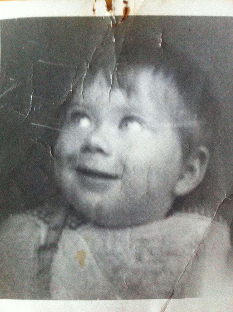 Monochrome photo of a chubby baby Bill looking up and to the viewer’s left. The photo is a bit screwed up and even has a rusty drawing pin mark where it must have once been pinned to a board. 