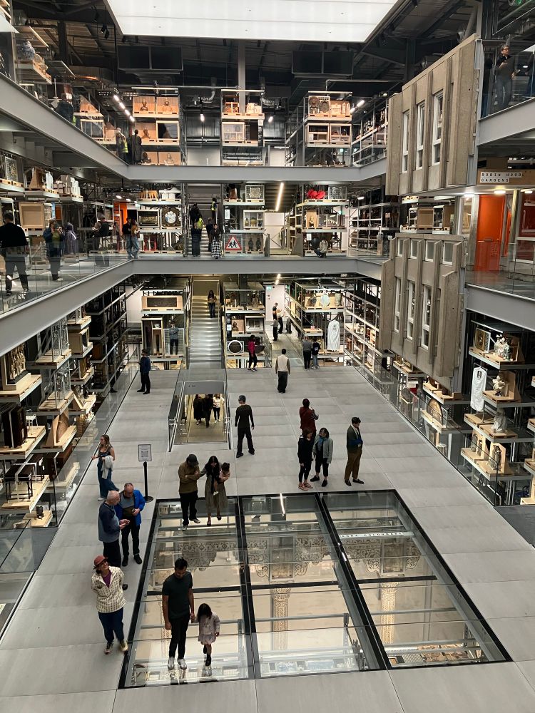 A view across a bright, modern atrium surrounded by 2 floors of museum pieces of warehouse style racking. People of all ages are moving slowly around, considering what they’re seeing.