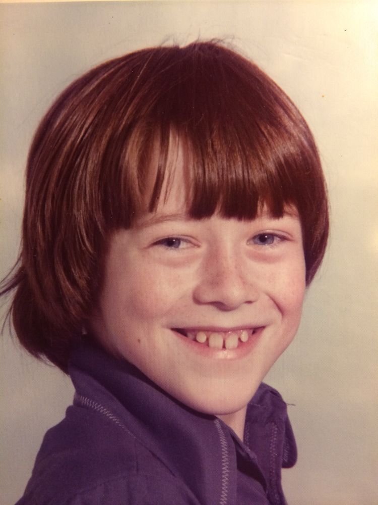 Bill is about 8 or 9, smiling towards the camera to his right. It’s a school style photo but he’s in his own 70s purple shirt. He has gappy teeth.