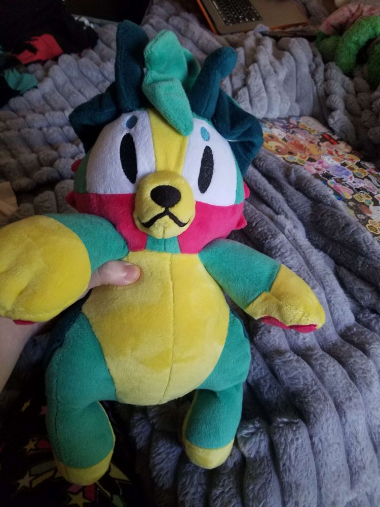 A teal puppy stuffed animal with a yellow belly and paws, pink cheeks, and dark green ears and antennae. It has white squares with embroidered eyes on its face, resembling glasses. It is being held up to the camera in front of a bed with a gray blanket, two computers, and some laundry in the background.