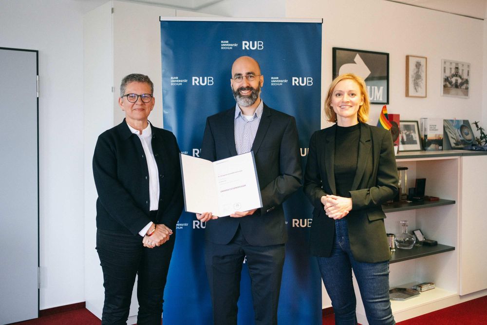 Three smiling people stand in front of a RUB banner. The man in the middle is holding a certificate. Photo credit: RUB, Kramer.