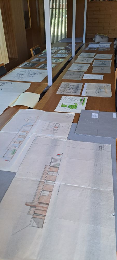 An assemblage of building plans and photographs laid out on the tables of an archive reading room.