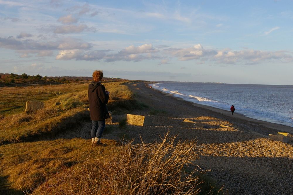 A view north along a curving coastline, in a slanting winter afternoon light. On the left are low dunes, on which the photographer is standing; occasional World War II anti-tank defences, in the form of concrete cubes, jut out at angles.  On the right of the picture the shadows of the dunes cover the beach, then beyond that the sea, coloured slightly purple by the setting sun.  A young man with red curly hair, wearing a duffle coat, is standing not far from the photographer, looking away up the coast; other figures are dotted along the beach at various intervals.
