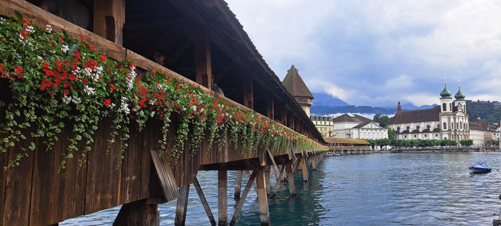 A long wooden covered bridge over a river, draped in red and white geraniums; a baroque church with two onion-domed towers is on the far side of the river.
