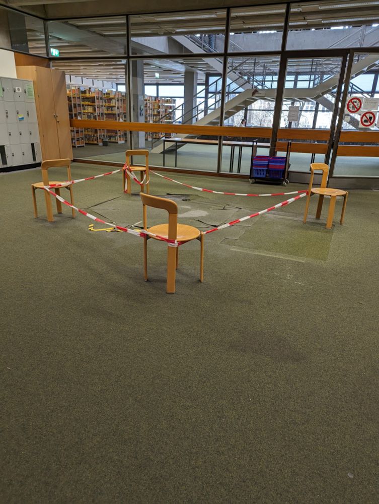 Das Foto zeigt zerbrochene Bodenplatten, die notdürftig mit Absperrband und vier Stühlen abgesichert wurden. Im Hintergrund ist eine Bibliothek sichtbar, das Foto wurde in einem Universitätsgebäude aufgenommen. 