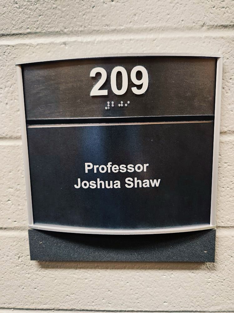 A photograph of black sign on a white wall which reads “Professor Joshua Shaw”.