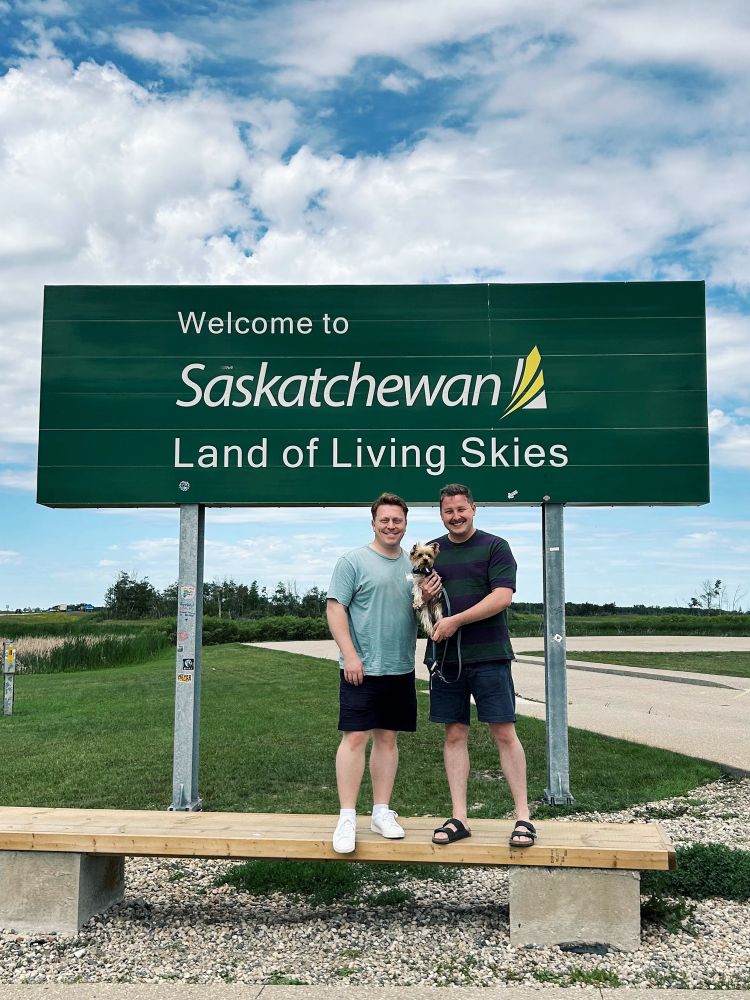 Two men and their dog stand in front of a sign which reads, “Welcome to Saskatchewan - Land of Living Skies”.