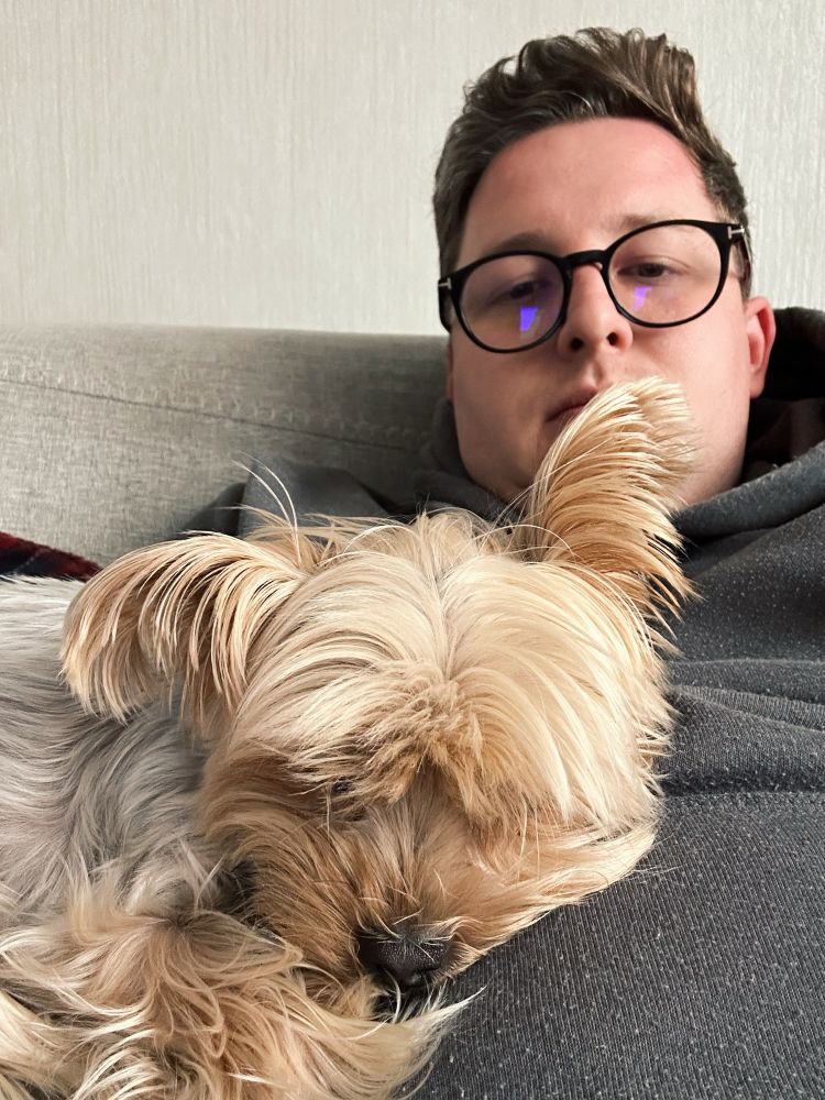 A human is seated in a reclined position on a couch, with a Yorkshire Terrier curled up, asleep on his stomach.