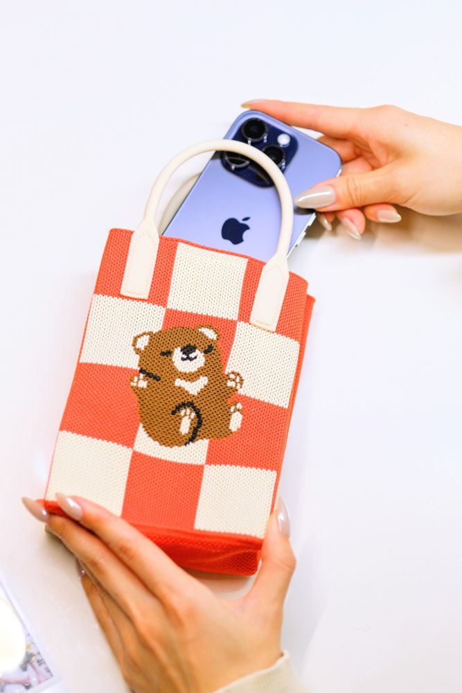 A photo of a knit bag being held with a purple iPhone being taken out of it. The knit bag features a design of a small brown bear lounging against a red and white checkered pattern.