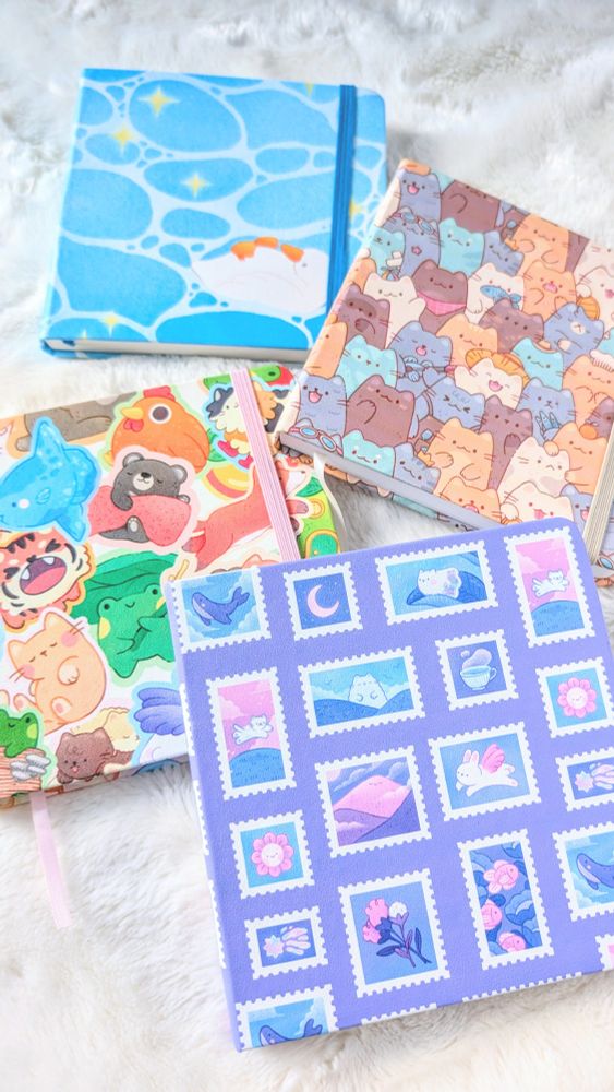 Photo of four square sketchbooks lying on a white blanket. The sketchbook cover designs are as follows: a) A series of stamps on a purple background. The stamps feature dreamy scenes and characters such as flowers and rabbits with wings, moons, a cup of tea. b) A collage of character stickers branding from bears in blankets, green frogs holding up a lily pad, foxes jumping, a blue mola mola. c) A pile of grey, white and brown cats as a pattern repeated over the cover. d) A small white duck floating on its back in blue waters.