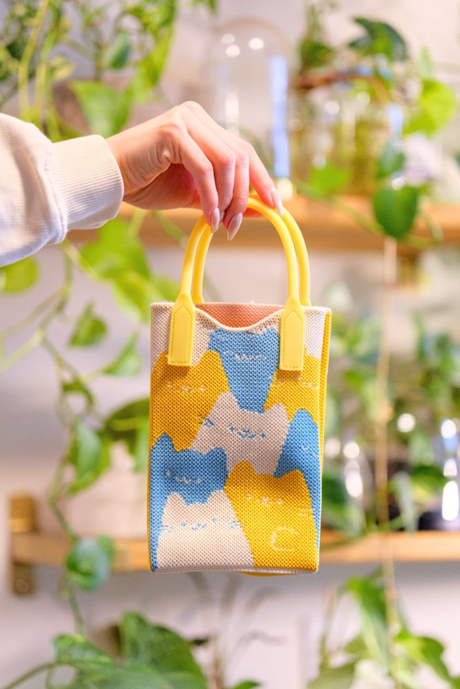 Photo of a knit bag being held against a backdrop of plants on shelves. The bag itself is comprised of a pattern of cats that are blue, white, and yellow.