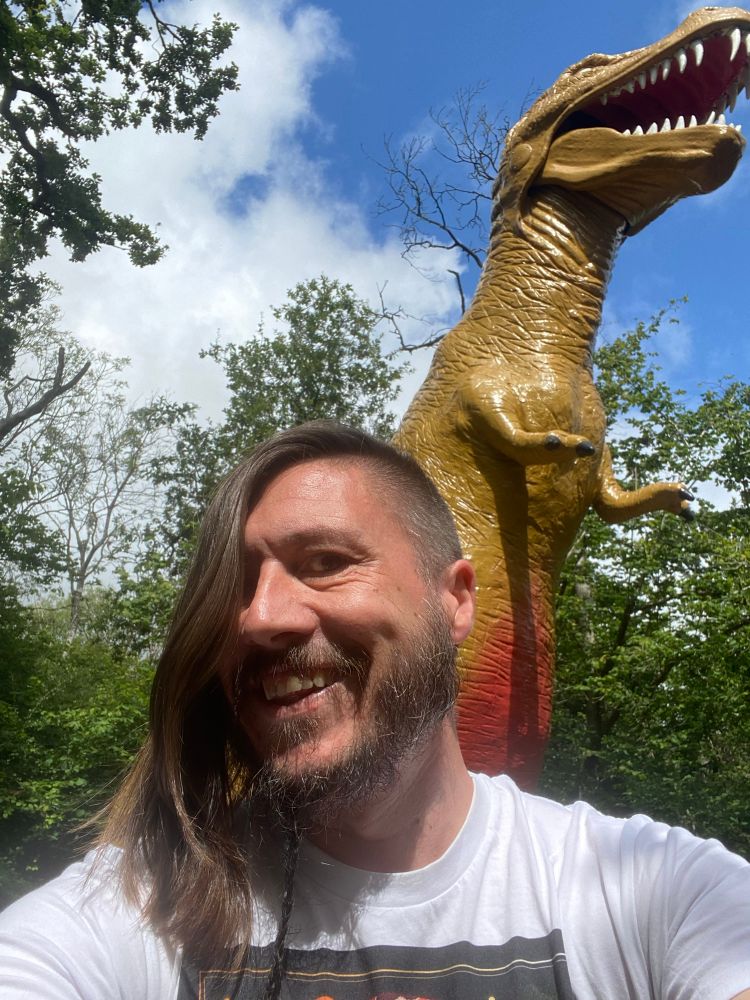 Smiling bearded man with dimples, standing in front of a tripodal Tyrannosaurus