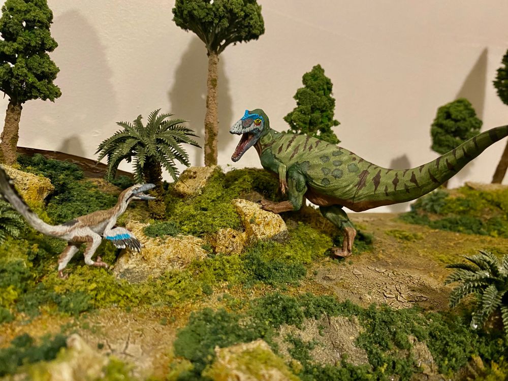 Green and brown theropod, with a flesh coloured chest, blue nasal crests and various stripes and mottled browns confronting a smaller feathered Theropod 