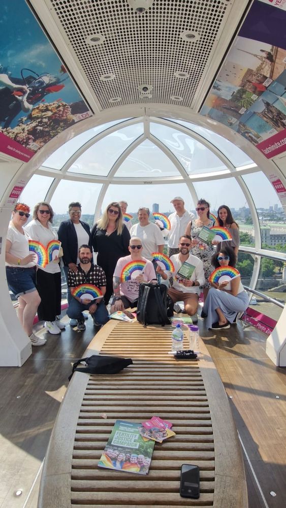 Group of people one of the London Eye pods. Everyone is smiling and wearing bright clothes, lots of people have rainbow fans.