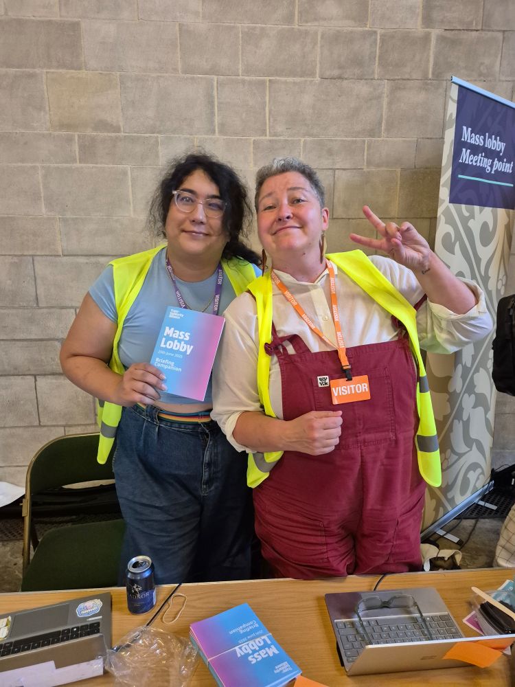 Two people cuddling side by side and smiling. Woman on the right is wearing hi vis over grey tshirt, jeans and rainbow belt and is holding up trans flag coloured leaflet for mass lobby. She has long wavy black hair in pony and is wearing glasses. Woman on right is in hi vis, red dungarees and has very short curly grey hair. 