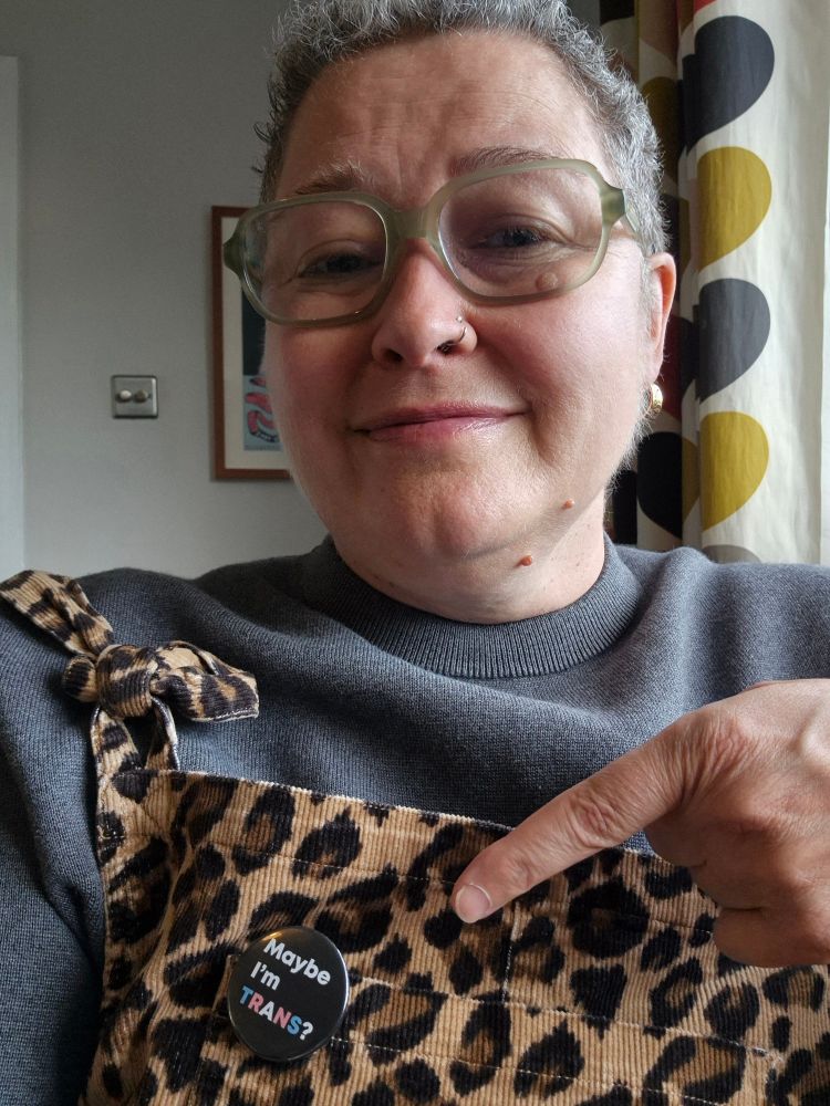 Picture of person with short curly great hair and green glasses wearing grey jumper under leopard print dungarees. She is pointing at a pin badge on her dungarees bib that reads "Maybe Im TRANS? In trans flag colours on a black background.