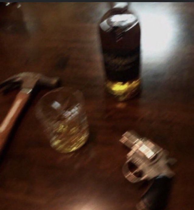 a blurry picture of a claw hammer, lowball glass full of whiskey, the bottle of whiskey, and a revolver, all sitting on a wooden table.