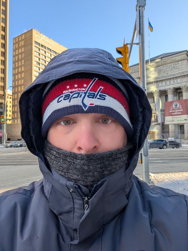 A selfie of a white man in a navy blue parka, with a red, white and blue Capitals beanie and a grey scarf wrapper around his face, standing in front of an intersection and office buildings.