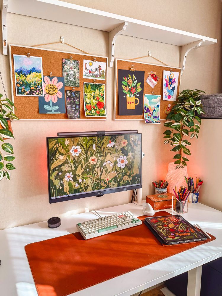 A photo of my desk with some sunlight pouring in. There is a monitor with a floral background, a brown desk mat, a beige keyboard, an iPad with a drawing on it, and some cups with pens in them.