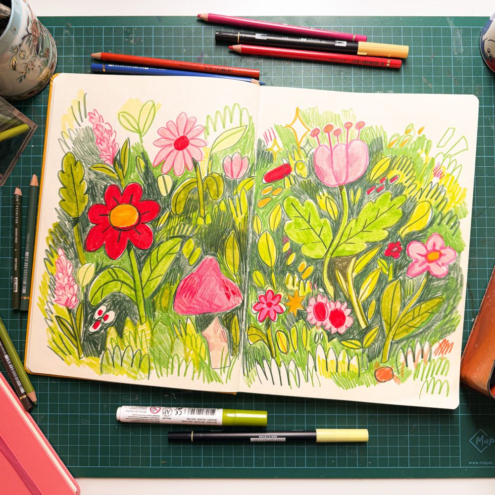 A photo of an A4 sketchbook open on a desk, inside of which is a very scribbly drawing of pink flowers and green leaves. Surrounding it are some scattered pens and pencils.