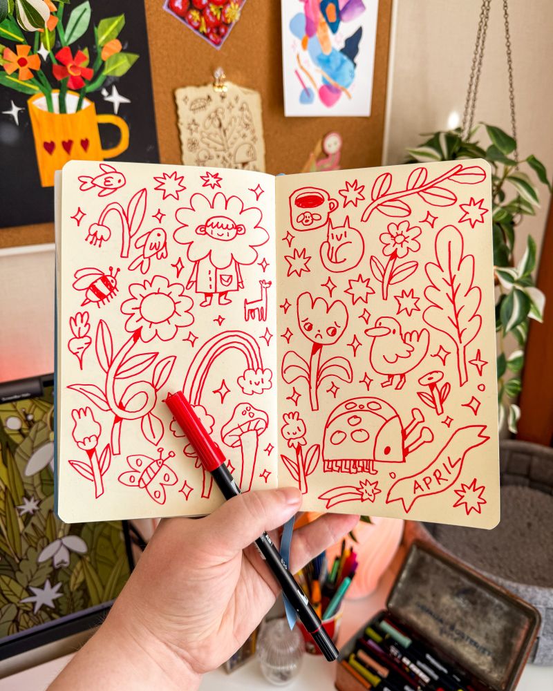 A photo of a sketchbook being held open in front of a desk, inside are doodles in red marker of different silly and cute items scattered on the page: a bee, fish, rainbow, flowers, ladybug, duck, cat etc. In the hand holding the sketchbook is also the red marker used for the spread.