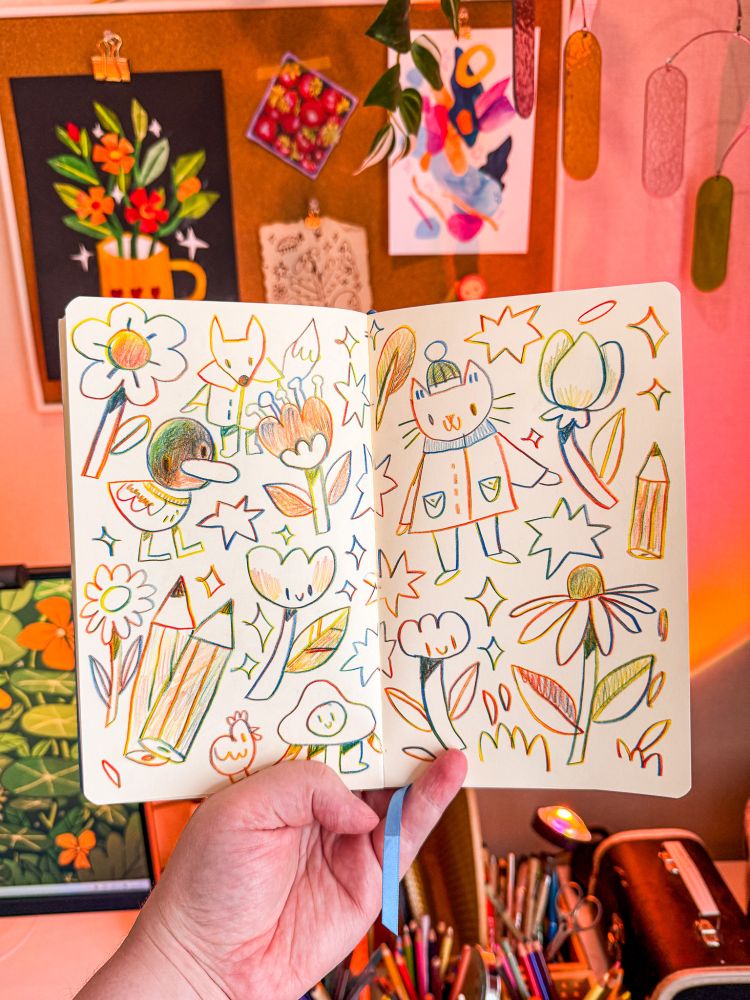 A photo of my hand holding up a sketchbook in front of my desk. Inside the sketchbook are lots of little drawings done with rainbow pencil, featuring a cat character, a duck, flowers, pencils and a fox. Behind the sketchbook is a warm pink light, a monitor and some pens and pencils in cups.