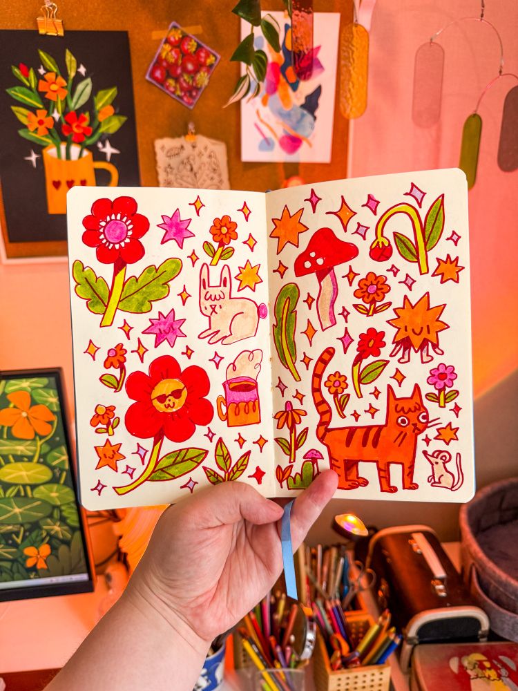 A photo of my sketchbook being held up in front of my desk. Inside the sketchbook are little drawn items scattered on the page, with flowers, a bunny, a cat, a star guy and a mushroom.