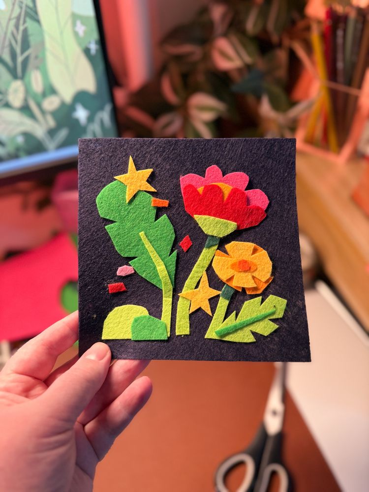 A photo of my hand holding a collage made out of felt. It features two flowers and some leaves made out of cut out felt, on a dark felt background! 
