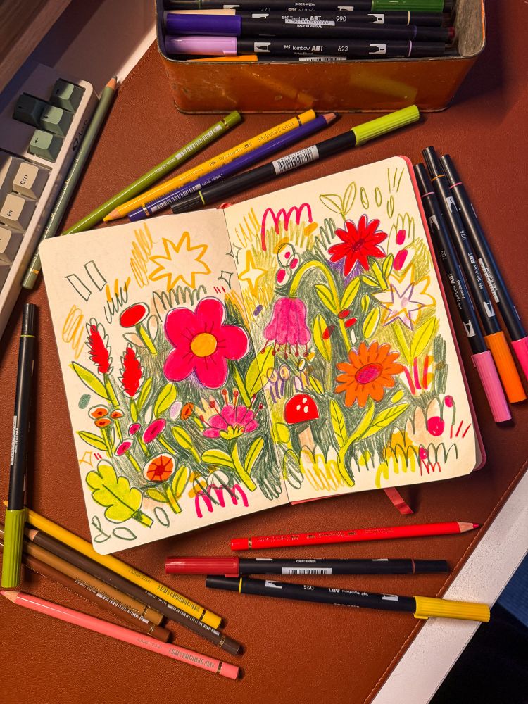 A photo of my sketchbook on my desk, with some scribbly flowers inside, and surrounded by pens and pencils.